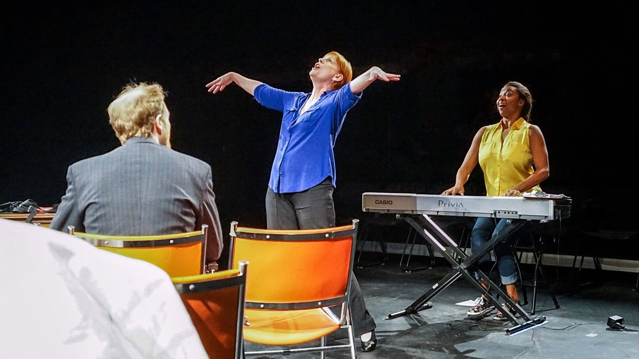 Three performers in a scene from The Final Say, with one standing center stage and another seated at a keyboard